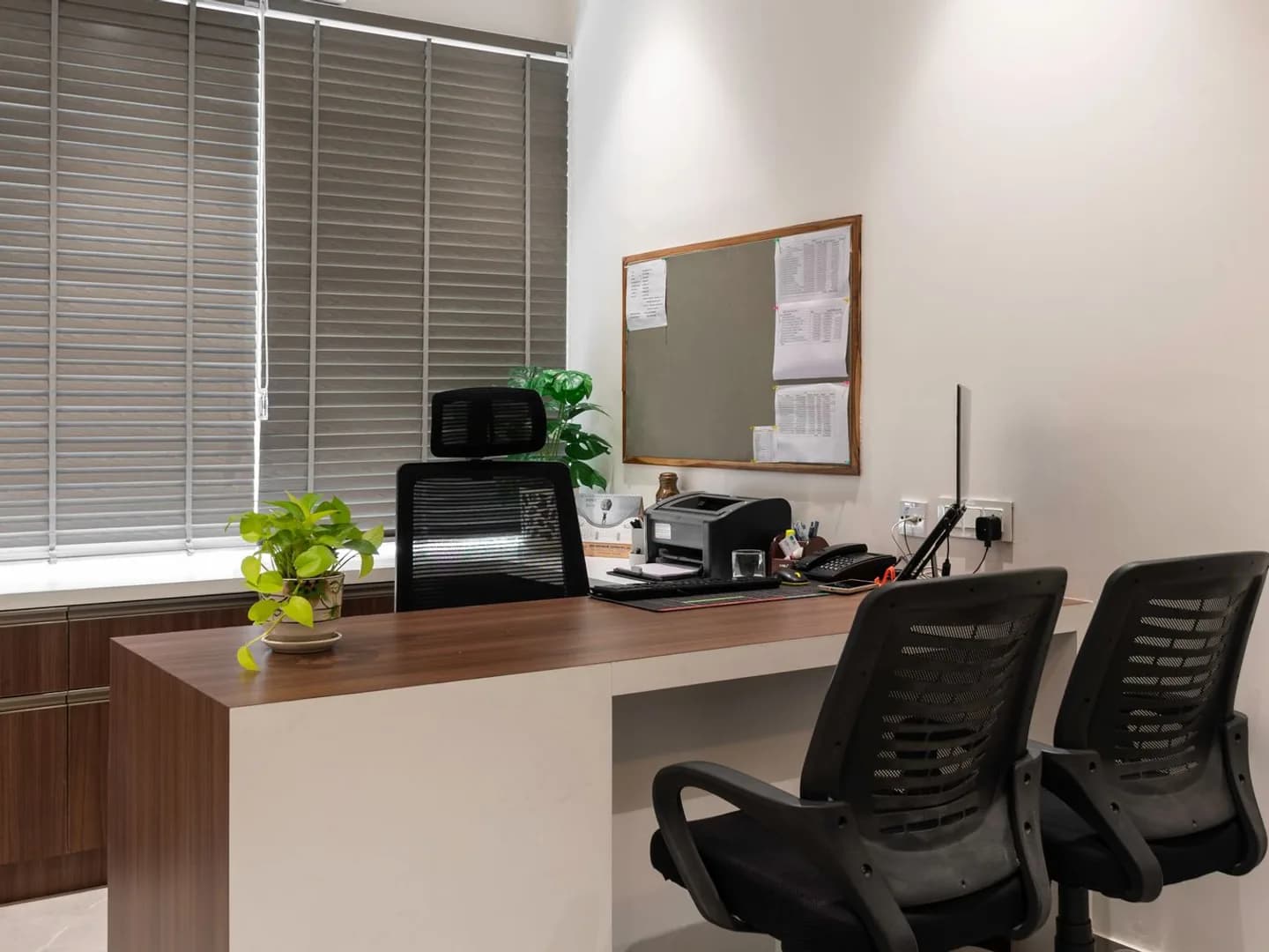 An empty office with a wooden desk, mesh chairs, window blinds and potted plants